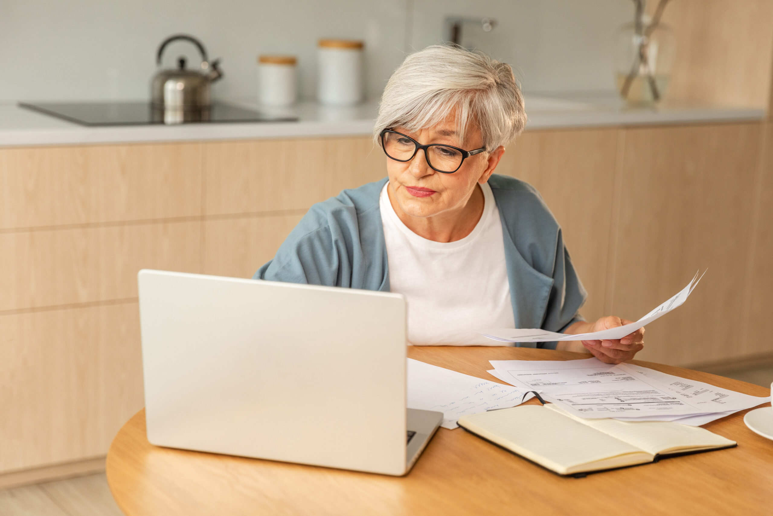 Middle aged senior woman sit with laptop and paper document, Pensive older mature lady reading paper bill pay online at home managing bank finances calculating taxes planning loan debt pension payment
